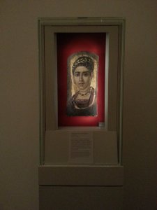 View of “Young Woman with Gilded Wreath” in Vitrine, Egypt, Roman Period (120–140 CE, encaustic with gold leaf on wood , 14⅜ x 7 in [36.5 x 17.8 cm]).  The Metropolitan Museum of Art, New York.  Photo © Deborah Feller.
